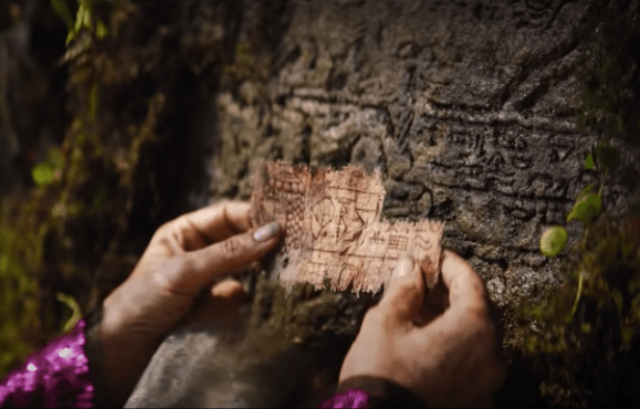 Two dirty hands holding up a scrap of papyrus with strange ideograms on , with a carved stone behind it with similar symbols.