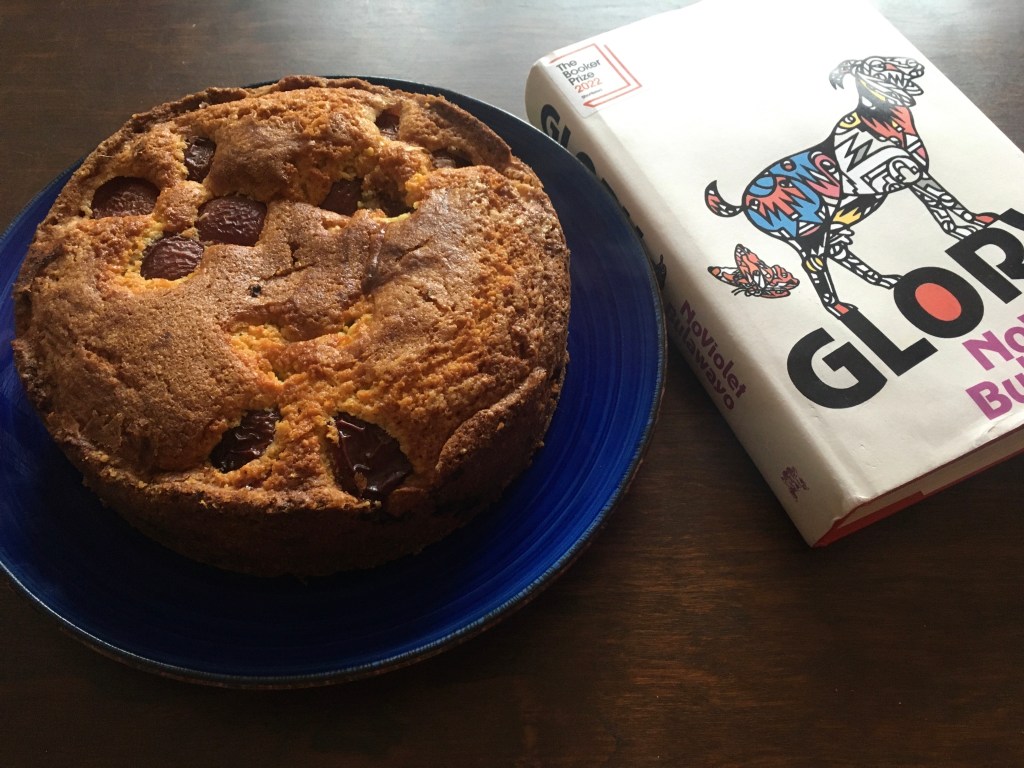 A round brown cake with baked plum quarters on the the top on a blue plate next to a copy of Glory by NoViolet Bulawayo