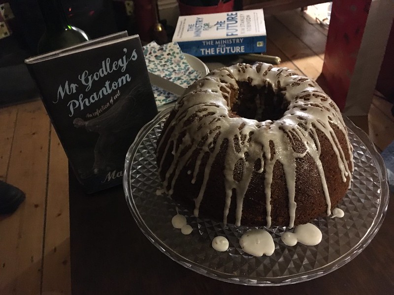 A copy of the book 'Mr Godley's Phantom' standing up with a gugulhupf cake with icing dripped down it on a cake stand in front of the book. At the back is a copy of 'The Ministry for the Future' by Kim Stanley Robinson