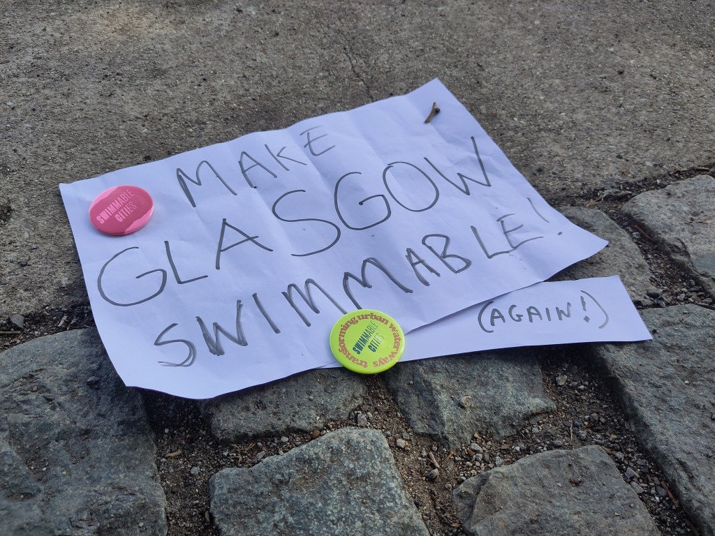 A handmade poster laid on quayside cobbles with the text 'Make Glasgow Swimmable' and the word 'Again!' on a smaller piece of paper attached at he bottom with a Swimmable Cities badge.